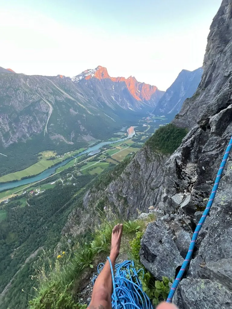 A climber's bare foot and coiled blue rope on a grassy ledge high above a valley river, with peaks lit orange