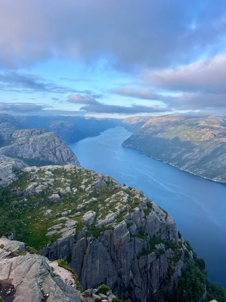 Looking down a long blue fjord from a high clifftop, steep grey rock walls on both sides under pink-tinged cloud