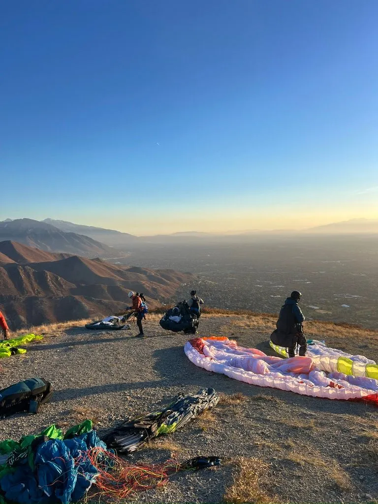 Pilots and spread-out wings on a gravel launch high above a hazy valley city, in low golden light