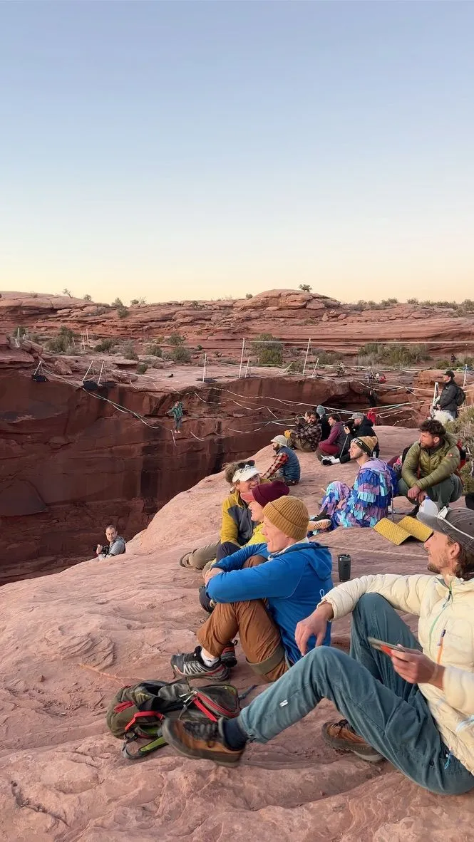 A crowd on a sandstone rim in low sun, watching a walker on one of several highlines across a red-rock gap
