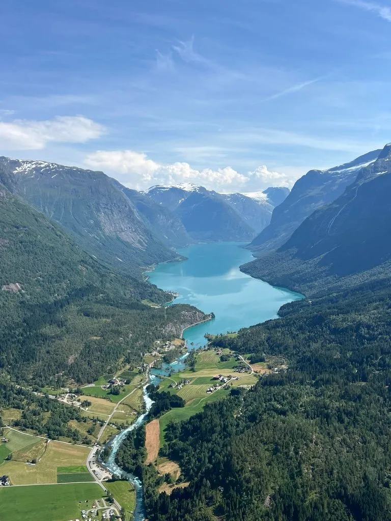 Looking down a green valley to a turquoise lake between steep mountains, snow on the peaks, farms and a river below