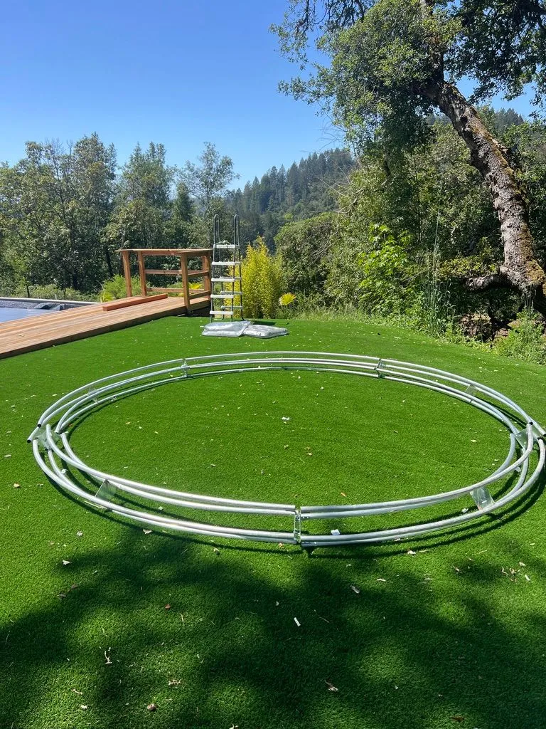 Four large concentric metal rings lying on artificial turf beside a wooden deck, with trees and wooded hills behind