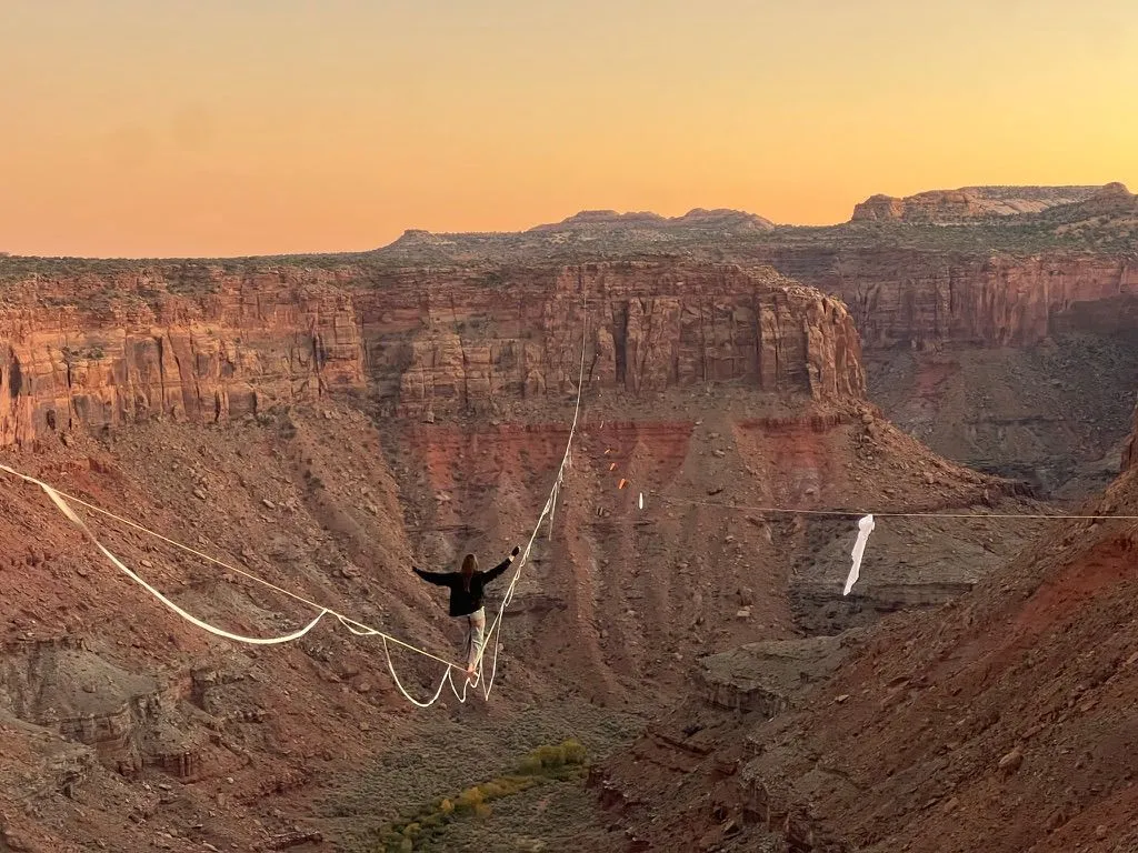 A highliner, arms out, walking a sagging line across a deep red-rock canyon under an orange sky