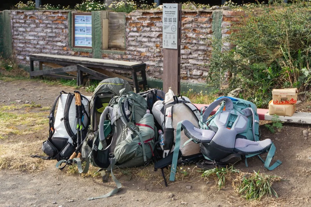 Loaded hiking backpacks and trekking poles piled on dry ground beside a bench, a brick wall and a park sign