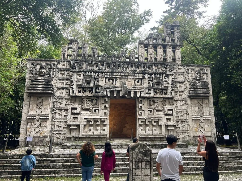 Visitors standing before a carved stone Maya-style temple facade with a dark central doorway, set among trees