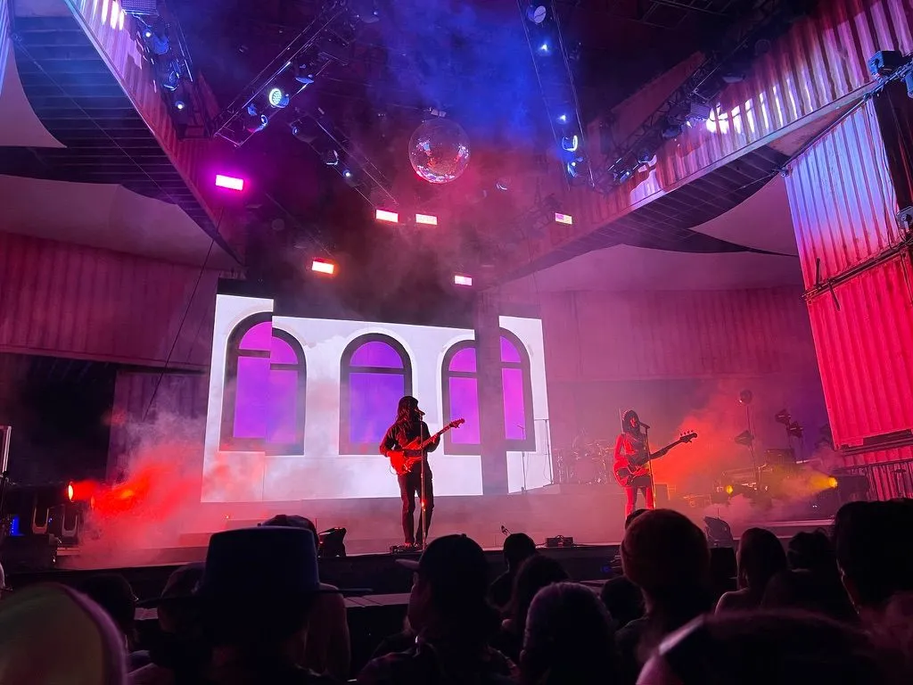 A band playing on a stage framed by stacked shipping containers, under a disco ball in red and purple haze