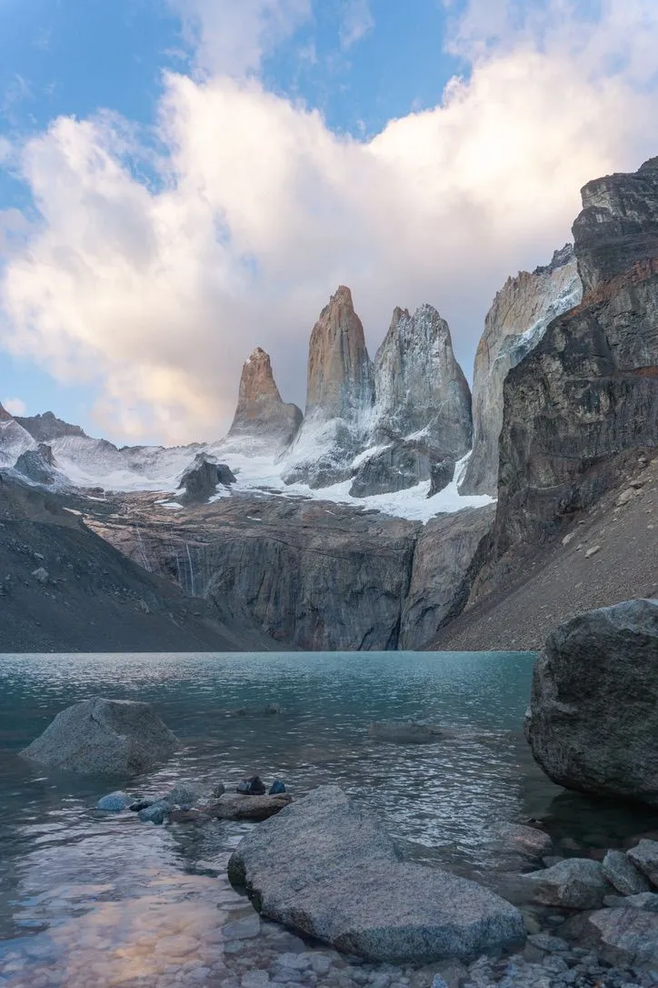 Three sheer granite towers rising above snowfields and a glacial lake, with boulders along the shore