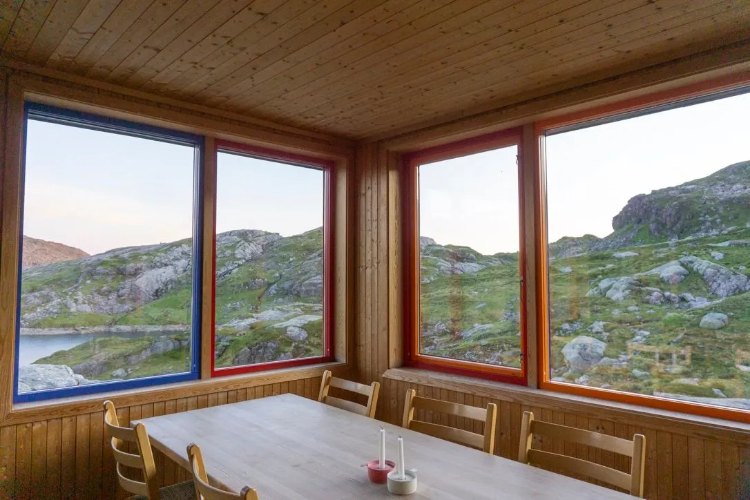 A long table in a wood-lined hut, its red, blue and orange framed windows looking onto rocky hills and a lake