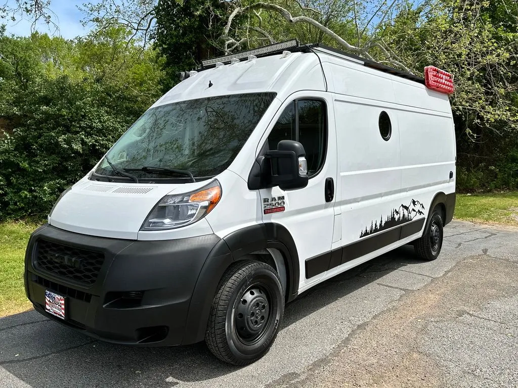 A white high-roof Ram ProMaster van with a round porthole window, roof rack, light bar and mountain decal