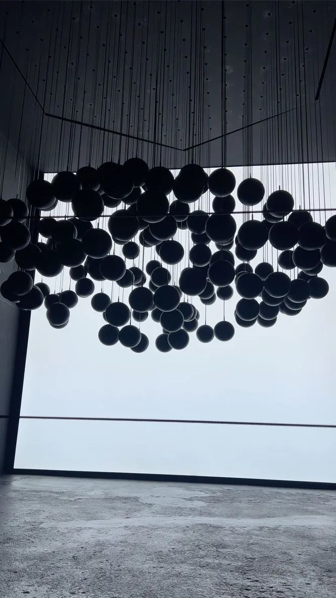 Hundreds of dark spheres hanging on thin cables from a high ceiling, silhouetted against a glowing white backdrop